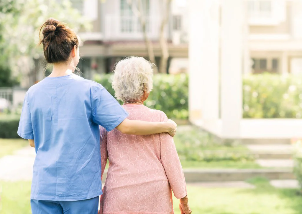 A care professional walking outdoors beside an older adult, offering gentle physical support, illustrating the type of care support and reassurance available in residential care settings