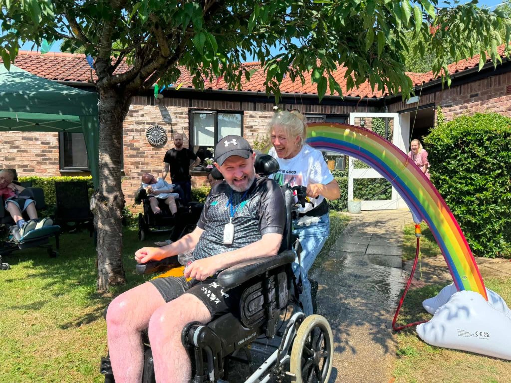 Resident at Meadow House being guided through a rainbow sprinkler wheelchair wash in the garden